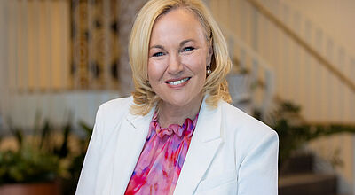 Professionelles Portrait einer Frau mit blonden, halblangen Haaren in weißem Blazer und pink-violetter Seidenbluse vor einem eleganten Treppenhaus-Hintergrund.