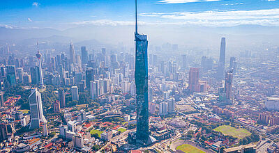 Blick auf die Skyline von Kuala Lumpur, Malaysia, mit dem dominierenden Merdeka 118 Wolkenkratzer im Zentrum, umgeben von zahlreichen Hochhäusern, sportlichen Anlagen und grünen Flächen bei klarem, blauem Himmel.