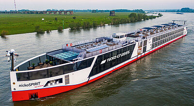Die Flussflotte von Nicko Cruises für 2026 umfasst 21 Schiffe, darunter die moderne Nicko Spirit Ein modernes Flusskreuzfahrtschiff mit senkrechtem rot-weißem Bug fährt durch eine weite Flussbiegung. Die Liegestühle auf dem Sonnendeck sind allesamt leer, am Ufer sind Wiesen zu sehen.