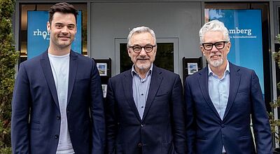 Die Geschäftsführung von Rhomberg Reisen (von links): Marco Wohlfahrt, Ioannis Afukatudis und Stefan Müller Drei Männer stehen lächelnd nebeneinander vor dem Eingang des Veranstalters „Rhomberg – Die Reisemaßschneiderei“. Sie tragen sportlich-elegante Outfits mit dunklen Blazern und Jeans.