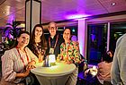 Ein Mann und drei Frauen an einem Stehtisch in einer Bar, die in lilanes Licht getaucht ist