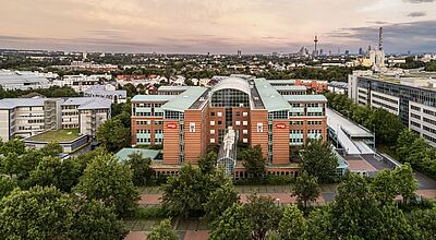 Luftaufnahme eines großen Bürogebäudes mit roter Backsteinfassade und Glasdach inmitten einer grünen Umgebung, im Hintergrund die Skyline von Frankfurt am Main mit dem Fernsehturm und weiteren Hochhäusern bei Sonnenuntergang.