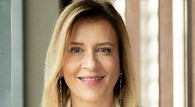 Eine Frau mittleren Alters mit langen blonden Haaren und braunen Augen lächelt in die Kamera. Sie trägt ein weißes Jackett.