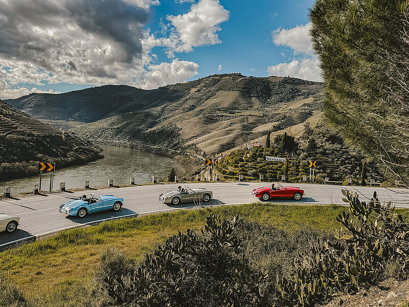Drei klassische Cabriolets fahren auf einer kurvenreichen Straße entlang eines Flusses durch eine malerische Hügellandschaft mit Weinbergen im Hintergrund. Ein Schild mit der Aufschrift „Graham’s“ deutet auf eine Weinkellerei hin. Der Himmel ist teils bewölkt, teils sonnig, und die Szene vermittelt ein Gefühl von Nostalgie und Reisefreude.