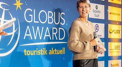 Verleihung des Globus Award von touristik aktuell – Gewinnerin mit kurzen Haaren und mit braunem Pullover auf der Bühne mit Auszeichnung und Mikrofon beim Branchenevent der Touristik“