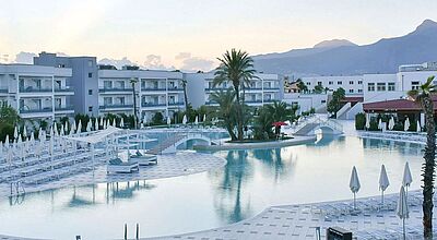 Großzügige Hotelanlage mit mehreren weißen Gebäuden, einem weitläufigen Poolbereich mit Liegen und Sonnenschirmen sowie Palmen im Vordergrund; im Hintergrund sind Berge unter klarem Himmel sichtbar