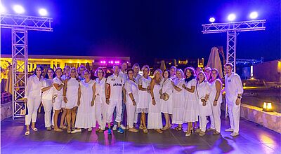 Eine große Gruppe von etwa zwanzig Personen steht abends im Freien unter blau-violetter Beleuchtung. Alle tragen weiße Kleidung, was auf eine White-Party oder ein sommerliches Gruppenevent hindeutet. Im Hintergrund sind Lichter, Gebäude und eine offene Terrasse sichtbar.