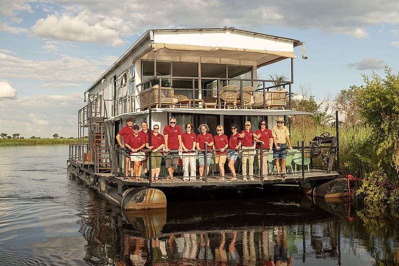Zweistöckiges Hausboot mit Aussichtsdeck auf dem Chobe-Fluss, ausgestattet mit Terrassen und Sitzgelegenheiten, zahlreiche Menschen in roten Hemden lehnen an der Reling und lächeln in die Kamera.