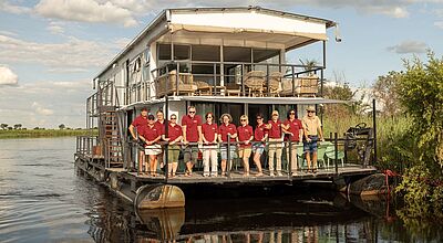 Zweistöckiges Hausboot mit Aussichtsdeck auf dem Chobe-Fluss, ausgestattet mit Terrassen und Sitzgelegenheiten, zahlreiche Menschen in roten Hemden lehnen an der Reling und lächeln in die Kamera.