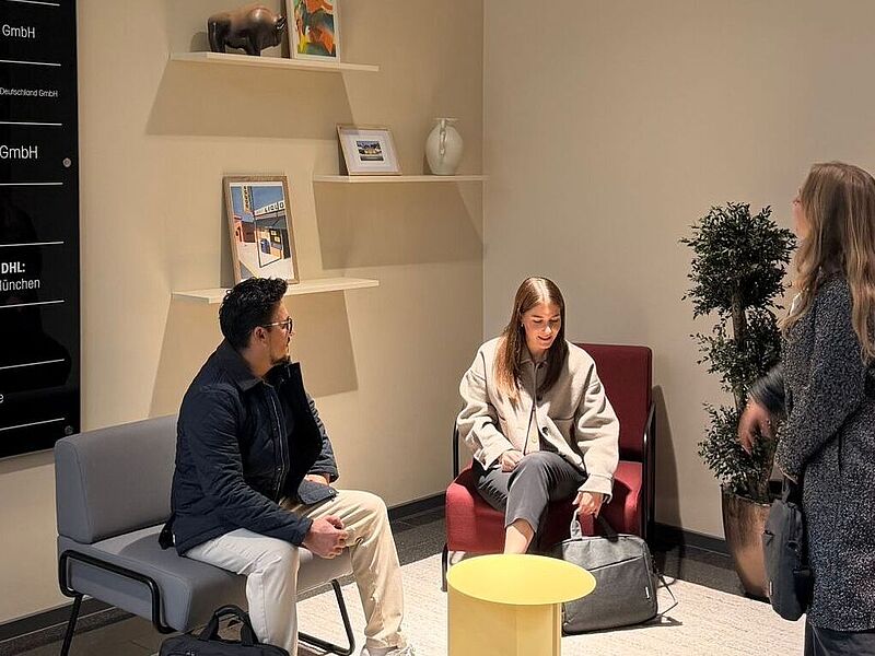 Insgesamt nutzen rund 40 Mitarbeitende die neuen Räumlichkeiten am Standort München – hier die Lobby. Drei Personen befinden sich in einer modernen Lobby. Zwei von ihnen sitzen auf Sesseln und unterhalten sich, während eine dritte Person im Mantel vorbeigeht. Hinter ihnen stehen ein Wandregal mit Deko-Objekten und ein schwarzes Firmenschild mit mehreren Unternehmensnamen. Im Vordergrund steht ein kleiner gelber Tisch.
