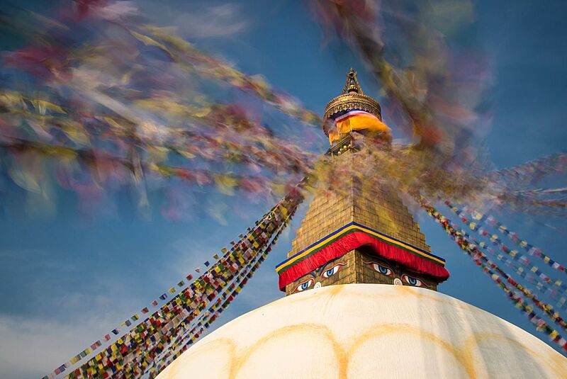 Ein großes buddhistisches Stupa mit goldener Turmspitze und den ikonischen, aufgemalten Buddha-Augen; bunte Gebetsfahnen wehen dynamisch im Wind vor einem klaren blauen Himmel und erzeugen verschwommene Farbstreifen im Bild.
