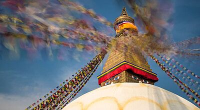 Vom 20. Februar bis zum 1. März können acht Reiseverkäufer mit Diamir Nepal erkunden Ein großes buddhistisches Stupa mit goldener Turmspitze und den ikonischen, aufgemalten Buddha-Augen; bunte Gebetsfahnen wehen dynamisch im Wind vor einem klaren blauen Himmel und erzeugen verschwommene Farbstreifen im Bild.