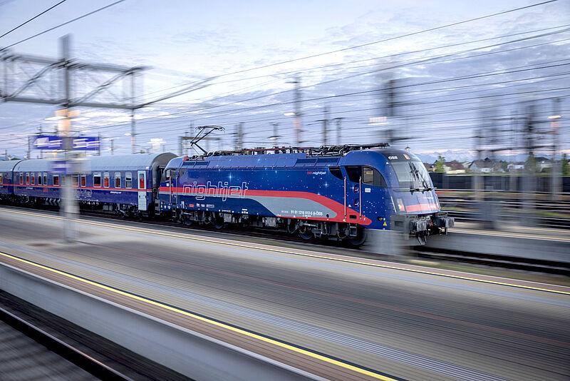 Ein Nachtzug der österreichischen Bahngesellschaft ÖBB in blauer, roter und grauer Lackierung fährt bei Dämmerung durch einen Bahnhof. Durch das Mitziehen der Kamera mit dem Zug ist die Umgebung verwischt, wodurch der Eindruck von Geschwindigkeit entsteht.