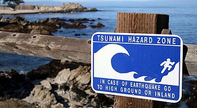 Blaues Tsunami-Warnschild an Holzgeländer vor felsiger Küstenlandschaft mit Hinweis auf Evakuierungsroute bei Erdbeben.