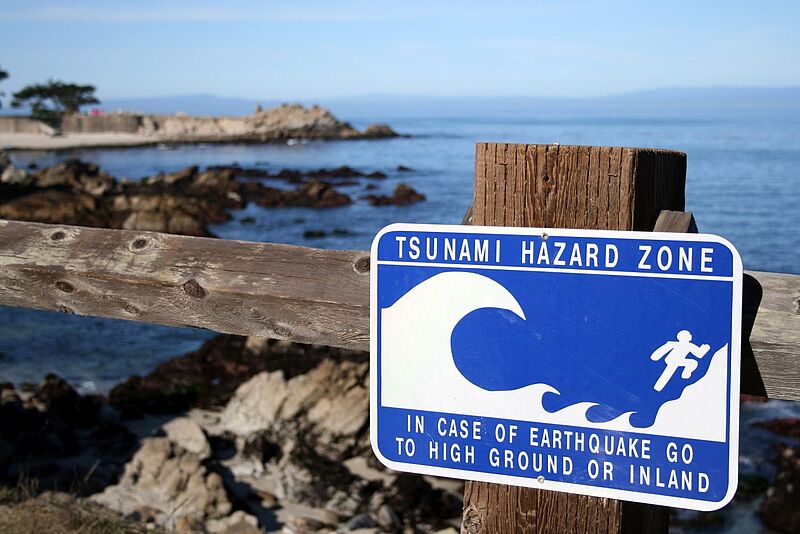 Blaues Tsunami-Warnschild an Holzgeländer vor felsiger Küstenlandschaft mit Hinweis auf Evakuierungsroute bei Erdbeben.