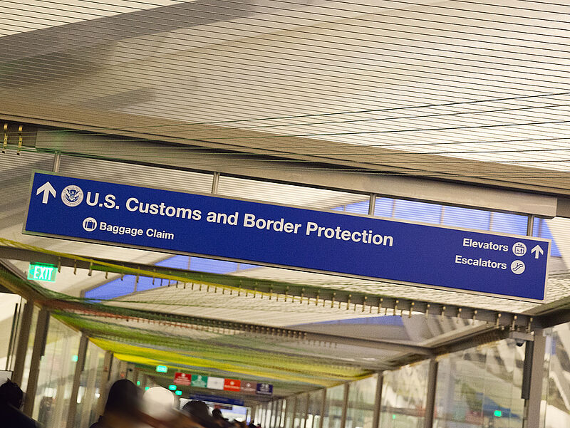 Blaues Hinweisschild der US Customs and Border Protection mit Richtungspfeilen zur Gepäckausgabe und Aufzügen/Rolltreppen an der Decke eines Flughafenterminals.