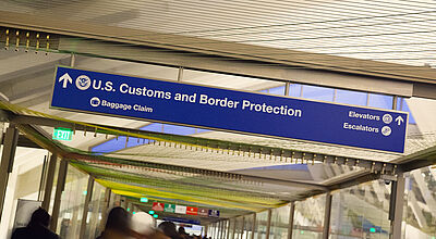 Blaues Hinweisschild der US Customs and Border Protection mit Richtungspfeilen zur Gepäckausgabe und Aufzügen/Rolltreppen an der Decke eines Flughafenterminals.