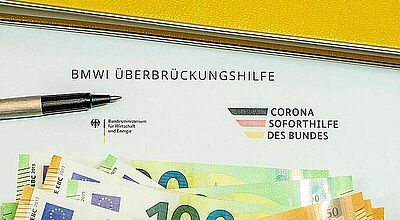 Die Schlussabrechnung der Überbrückungshilfe bereiten vielen Reisebüros nach wie vor Sorgen. Dokument zur BMWi-Überbrückungshilfe und Corona-Soforthilfe des Bundes, daneben ein Kugelschreiber und mehrere Euro-Geldscheine.