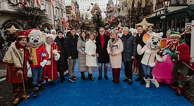 Foto von einer größeren Gruppe von Menschen, die auf einem blauen Teppich in einem Vergnügungspark stehen. Zwei der abgebildeten Personen sind als Mäuse verkleidet, drei weitere tragen ebenfalls Kostüme. Die anderen sind normal gekleidet.