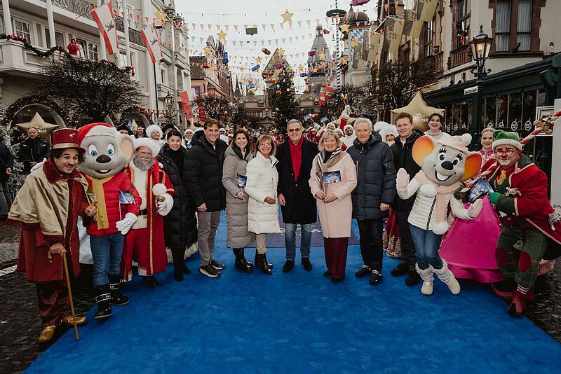 Foto von einer größeren Gruppe von Menschen, die auf einem blauen Teppich in einem Vergnügungspark stehen. Zwei der abgebildeten Personen sind als Mäuse verkleidet, drei weitere tragen ebenfalls Kostüme. Die anderen sind normal gekleidet.
