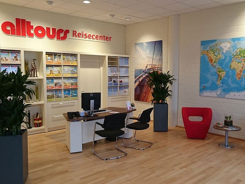 Das Bild zeigt das Innere eines modernen Reisebüros mit dem Namen "alltours Reisecenter". Im Vordergrund steht ein Empfangstisch mit Computer und zwei schwarzen Stühlen. Rechts davon befinden sich zwei rote Designerstühle und ein kleiner Tisch mit Pflanzen und Prospekten. Im Hintergrund sieht man Regale mit Reisekatalogen, dekorative Pflanzen, ein großes Wandbild von einem Segelboot auf dem Meer und eine Weltkarte an der Wand. Der Raum ist hell, freundlich und aufgeräumt gestaltet