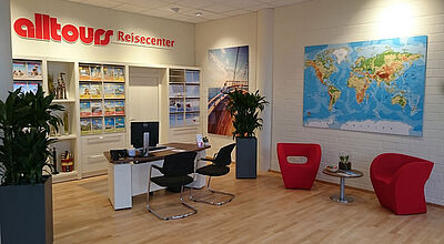 Das Bild zeigt das Innere eines modernen Reisebüros mit dem Namen "alltours Reisecenter". Im Vordergrund steht ein Empfangstisch mit Computer und zwei schwarzen Stühlen. Rechts davon befinden sich zwei rote Designerstühle und ein kleiner Tisch mit Pflanzen und Prospekten. Im Hintergrund sieht man Regale mit Reisekatalogen, dekorative Pflanzen, ein großes Wandbild von einem Segelboot auf dem Meer und eine Weltkarte an der Wand. Der Raum ist hell, freundlich und aufgeräumt gestaltet