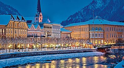 Winterliche Stadtansicht mit verschneiten Häusern, einer Kirche mit beleuchtetem Kirchturm und bunten Fassaden entlang eines Flusses. Die Uferpromenade ist festlich beleuchtet, im Hintergrund erheben sich schneebedeckte Berge.