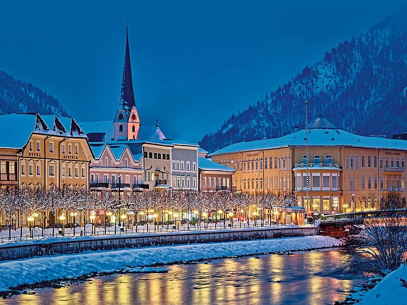 Winterliche Stadtansicht mit verschneiten Häusern, einer Kirche mit beleuchtetem Kirchturm und bunten Fassaden entlang eines Flusses. Die Uferpromenade ist festlich beleuchtet, im Hintergrund erheben sich schneebedeckte Berge.