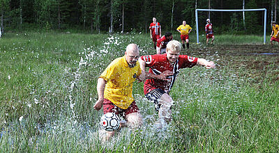 Männer in roten und gelben Trikots spielen auf einer matschigen Wiese mit hohem Gras und Pfützen Fußball