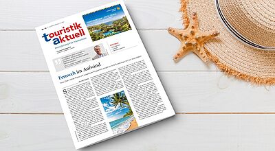 Eine Ausgabe der Fachzeitung „touristik aktuell“ liegt auf einem weißen Holztisch. Daneben befinden sich ein Strohhut mit schwarzem Band und eine orangefarbene Seestern-Dekoration. Die Titelseite zeigt ein tropisches Reiseziel mit Palmen, Pool und Strand sowie eine Schlagzeile „Fernweh im Aufwind“.