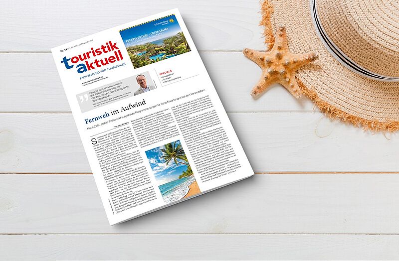 Eine Ausgabe der Fachzeitung „touristik aktuell“ liegt auf einem weißen Holztisch. Daneben befinden sich ein Strohhut mit schwarzem Band und eine orangefarbene Seestern-Dekoration. Die Titelseite zeigt ein tropisches Reiseziel mit Palmen, Pool und Strand sowie eine Schlagzeile „Fernweh im Aufwind“.