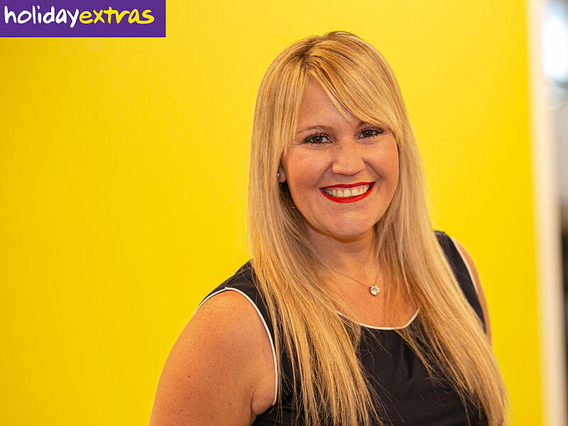 Eine Frau mit langen blonden Haaren und dunkelblauem ärmellosen Oberteil steht vor einer gelben Wand. Oben links im Bild befindet sich das Logo "holidayextras" auf lila Hintergrund. Im Hintergrund rechts sind unscharf Büromöbel und Fenster zu erkennen.