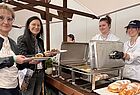 Zwei Frauen mit Tellern mit Speisen in der Hand an einem Essensstand, dahinter zwei Frauen in weißer Kleidung, die Essen austeilen