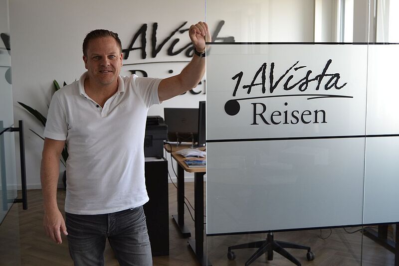 Geschäftsführer Ralph Dombrowski am Unternehmenssitz von 1A Vista Reisen in Köln Ein Mann mittleren Alters in weißem Poloshirt posiert vor seinem modernen Büro und hat den Arm lässig an eine Glastür gelehnt.