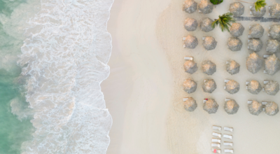 Luftaufnahme von Karibik-Strand mit Palmen, türkisblauem Meer, weißen Wellen und Strohhütten-Sonnenschirmen tropisches Urlaubsziel für Strandurlaub und Erholung.