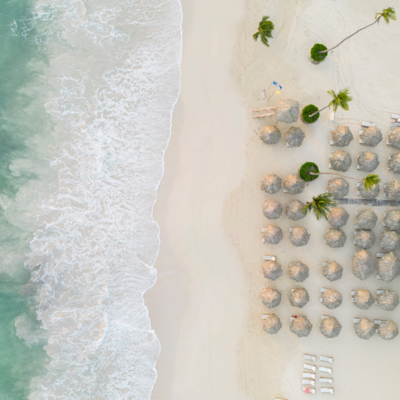 Luftaufnahme von Karibik-Strand mit Palmen, türkisblauem Meer, weißen Wellen und Strohhütten-Sonnenschirmen tropisches Urlaubsziel für Strandurlaub und Erholung.