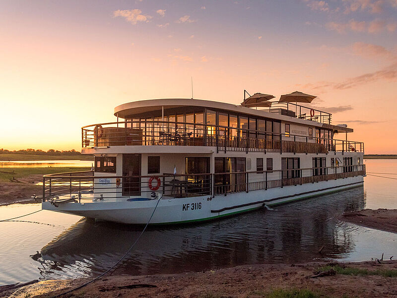 Auf dem Sambesi in Afrika liegt nur wenige Meter vom flachen, sandigen Ufer ein Boutique-Flusskreuzfahrtschiff mit zwei Decks. Durch die gläserne Lounge auf dem Oberdeck scheint die untergehende Abendsonne.