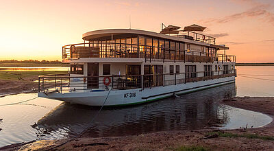 Auf dem Sambesi in Afrika liegt nur wenige Meter vom flachen, sandigen Ufer ein Boutique-Flusskreuzfahrtschiff mit zwei Decks. Durch die gläserne Lounge auf dem Oberdeck scheint die untergehende Abendsonne.