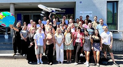 Gruppenfoto vor dem Gebäude der Bethmannschule Frankfurt. Etwa 35 junge Erwachsene, vermutlich Auszubildende im Bereich Tourismus, stehen lächelnd in mehreren Reihen. Im Hintergrund ist das Schulgebäude mit einem Schild "Bethmannschule" zu sehen. Zwei Personen halten symbolisch einen aufblasbaren Globus und ein Flugzeugmodell hoch, was auf das Thema Reisen oder Tourismus hinweist. Es ist sonnig, die Stimmung wirkt fröhlich und sommerlich.