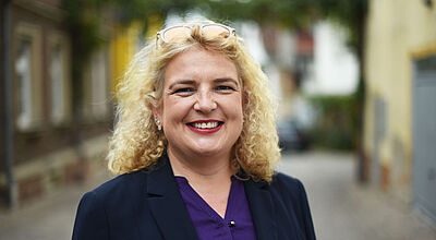 Eine blond gefärbte Frau mittleren Alters im dunklen Jackett und blauer Bluse steht vor einem unscharfen Hintergrund einer Hofeinfahrt