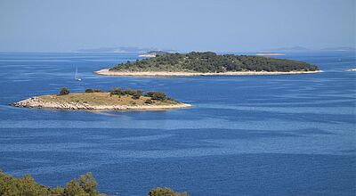 Im neuen Reiseperlen-Magazin werden auch Segelreisen nach Kroatien vorgestellt Blick vom Festland auf zwei kleine Karstinseln mit spärlichem Bewuchs. Das Meer ist tiefblau, zwischen den Inseln ist ein Segelboot zu sehen