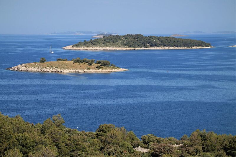 Im neuen Reiseperlen-Magazin werden auch Segelreisen nach Kroatien vorgestellt Blick vom Festland auf zwei kleine Karstinseln mit spärlichem Bewuchs. Das Meer ist tiefblau, zwischen den Inseln ist ein Segelboot zu sehen