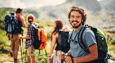 Der Katalog soll vor allem junge Reisende ansprechen, die in einer internationalen Gruppe unterwegs sein wollen. Foto: Todor Tsvetkov/iStock Eine Gruppe von Wandernden auf einem Bergpfad; im Vordergrund steht ein junger Mann mit Rucksack und Trekkingstöcken, der lächelnd in die Kamera blickt