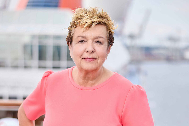 Reederei-Managerin Anfang sechzig mit kurzen braunen Haaren lehnt in lachsfarbenem Kleid an der Reling eines Kreuzfahrtschiffs und blickt freundlich in die Kamera