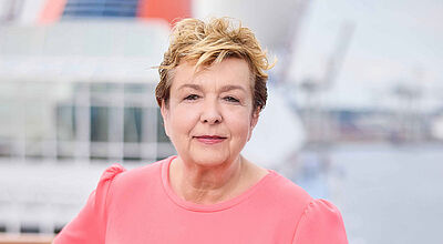 Seit Sommer am Ruder bei Hapag-Lloyd Cruises: Managing Director Isolde Susset Reederei-Managerin Anfang sechzig mit kurzen braunen Haaren lehnt in lachsfarbenem Kleid an der Reling eines Kreuzfahrtschiffs und blickt freundlich in die Kamera