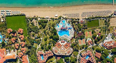 Ein Prozent Zusatzprovision gibt es unter anderem für den Verkauf von Reisen ins Asteria Family Resort in Belek Luftaufnahme eines großen Strandresorts an der türkisen Küste. In der Mitte befindet sich ein großes Schwimmbecken mit Wasserrutschen, umgeben von Liegestühlen und Palmen. Direkt darüber liegt ein Sandstrand mit vielen Sonnenschirmen und dem Meer. Rund um die Poolanlage verteilen sich Tennisplätze, grüne Rasenflächen, Wege und mehrere Gebäude mit roten Dächern. Unten im Bild sind weitere Hotelgebäude, Gärten und kleinere Pools zu sehen.