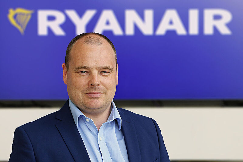 Ein Manager der Fluggesellschaft Ryanair steht mit hellblauem Hemd und dunkelblauem Jackett vor einem unscharfen Logo und Schriftzug von Ryanair im Hintergrund.