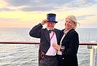Ein elegant gekleideter Mann mit Zylinder und eine elegant gekleidete Frau stehen an der Reling. Der Mann stellt ein RB aus Plastik auf seinen Zylinder