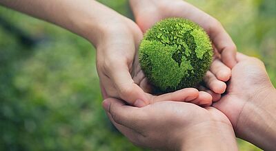 Mehrere Hände halten gemeinsam eine grüne, moosbedeckte Kugel in Form der Erde – ein Symbol für Umweltschutz, Nachhaltigkeit und gemeinschaftliche Verantwortung für unseren Planeten.“