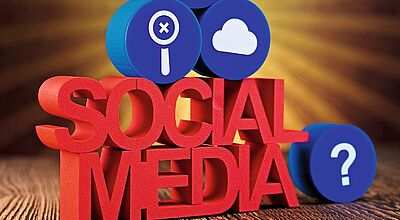 3D-Schriftzug "SOCIAL MEDIA" mit Symbolen für Geld, Suche, Cloud und Fragezeichen auf Holzuntergrund mit Strahlenkranz.